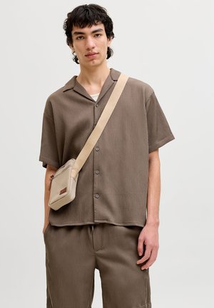 Νέος άνδρας που φοράει καφέ πουκάμισο με κοντά μανίκια και κουμπιά, καθώς και παντελόνι στο ίδιο χρώμα, κρατώντας μια μπεζ τσάντα χιαστί σε απλό φόντο.