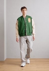 Chaqueta varsity verde y crema con letras amarillas, botones y parches en los hombros, combinada con jeans gris claro y zapatillas blancas.
