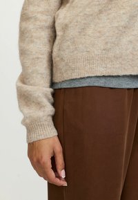 Beżowy sweter wełniany z prążkowanymi mankietami, noszony na szarym topie, w połączeniu z brązowymi zwężającymi się spodniami. Miękka tekstura i luźny krój.