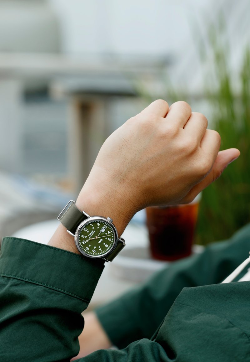 Grüne Militäruhr mit rundem Zifferblatt, weißen Zahlen und grauem Armband, getragen an einem Handgelenk mit grünem Hemd.