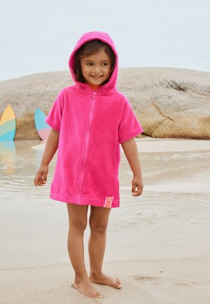 Enfant souriant en robe serviette à capuche rose, debout pieds nus sur du sable mouillé près de planches de surf colorées et d'un gros rocher.