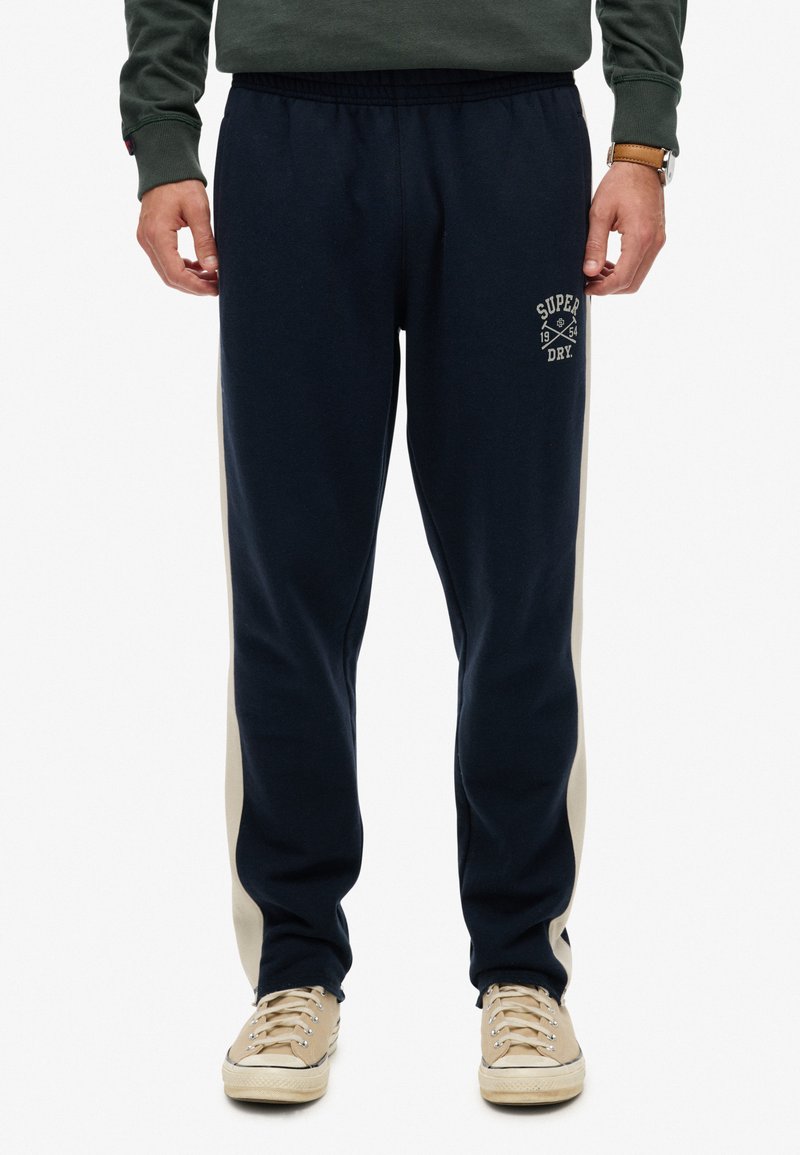 Marineblauwe joggingbroek gemaakt van zacht materiaal met een witte zijpaneel en geborduurd logo-detail. Beschikt over een elastische tailleband en een tapse pasvorm.