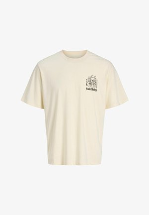 Nastolatek z kręconymi włosami uśmiecha się, ubrany w kremowy t-shirt z grafiką Palermo, szerokie niebieskie dżinsy i brązowe buty, stoi na białym tle.