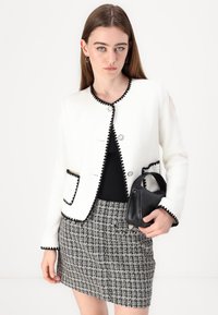 Veste blanche texturée avec un bord noir à volants, des boutons et des détails de poche, portée sur un haut noir et associée à une jupe à carreaux noir et blanc.