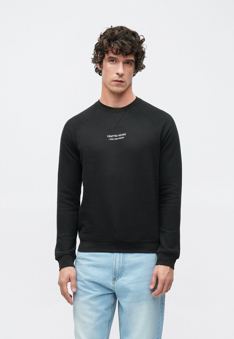 Musta pitkähihaisen collegepaita, jossa on resorit hihansuissa ja pyöreä pääntie. Rinnassa keskitetty valkoinen teksti "CRAFTED GOODS". Yhdistetty vaaleansinisten farkkujen kanssa.