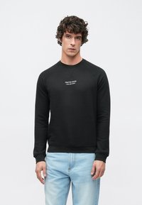 Sweat-shirt noir à manches longues, avec des poignets côtelés et un col rond. Texte blanc centré sur la poitrine avec l'inscription "CRAFTED GOODS". Associé à un jean bleu clair.