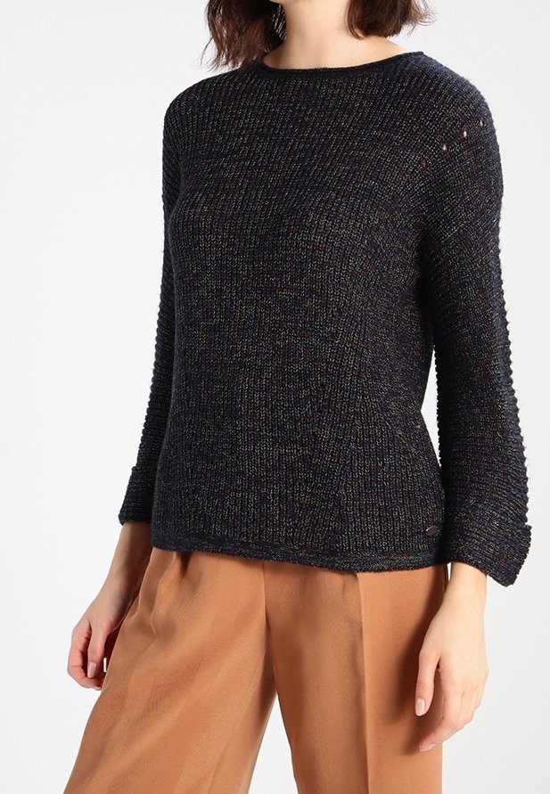 Femme portant un pull en maille noire texturée à manches longues et un pantalon marron ample, debout devant un fond blanc uni.