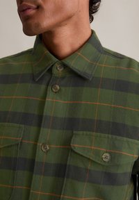 Πράσινο καρό πουκάμισο από υφασμένο ύφασμα, με δύο προσθετές τσέπες, κουμπιά μπροστά και πορτοκαλί λεπτομέρειες. Έχει γιακά και κλασική εφαρμογή.