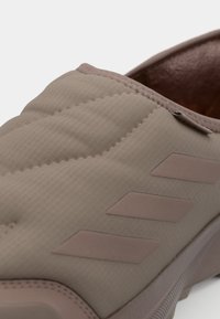 Chaussure slip-on rembourrée beige en matériau texturé, avec trois rayures beige foncé sur le côté et une semelle en caoutchouc lisse.