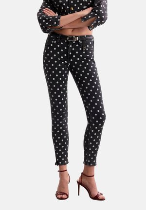 Femme portant un jean skinny taille haute noir et un chemisier court assorti à pois blancs, ainsi que des sandales à talons hauts bordeaux.