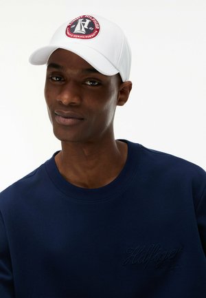 Hombre de piel oscura con una gorra blanca Tommy Hilfiger y una sudadera azul marino con el logo de Hilfiger bordado.