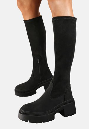 Bottes en suède noires jusqu'au genou avec une semelle en caoutchouc épaisse et une fermeture éclair sur le côté. Présente une texture lisse et un design minimaliste.