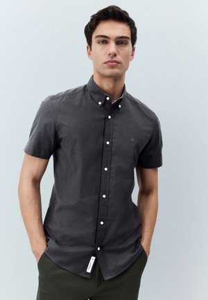 Mladi muškarac nosi usku tamnosivu košulju kratkih rukava s ovratnikom, stoji s rukama u džepovima na jednobojnoj pozadini.