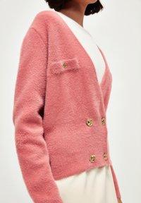 Femme portant un cardigan en peluche rose doux avec des boutons dorés sur un haut blanc, montrée de mi-torse à cou sur fond blanc.