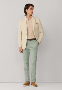 Homme debout portant un blazer beige avec un mouchoir de poche à motifs, un polo marron clair, un pantalon vert menthe, une ceinture marron et des chaussures en daim beige.