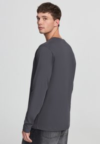 Maglietta grigia a maniche lunghe in cotone misto, con scollatura rotonda e vestibilità rilassata, indossata con jeans scuri.
