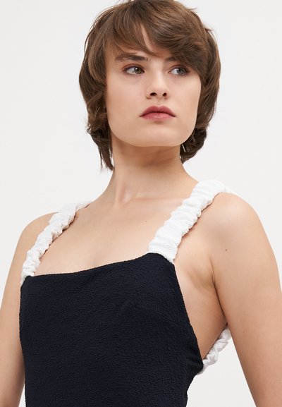Black dress with textured fabric, featuring white, gathered straps. Square neckline and sleeveless design highlight the shoulders.