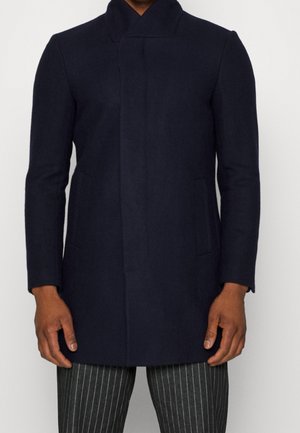 Homme portant un manteau bleu marine ajusté avec des boutons avant dissimulés et un pantalon à fines rayures noires, sur un fond blanc uni.