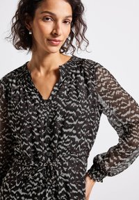 Femme aux cheveux bouclés portant un blouse à manches longues noire et blanche à motifs avec des boutons et une ceinture à la taille, posant devant un fond blanc.