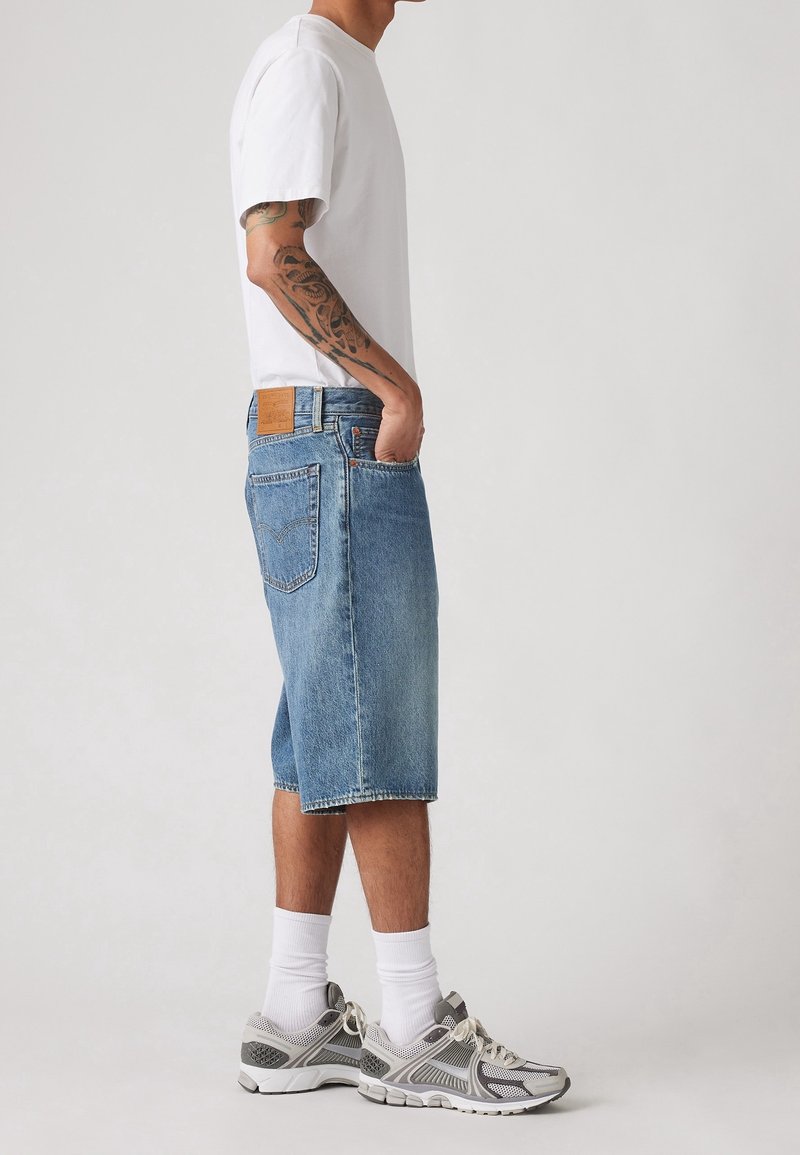 Homme portant un t-shirt blanc, un long short en jean bleu, des chaussettes blanches et des baskets grises, debout de côté les mains dans les poches.
