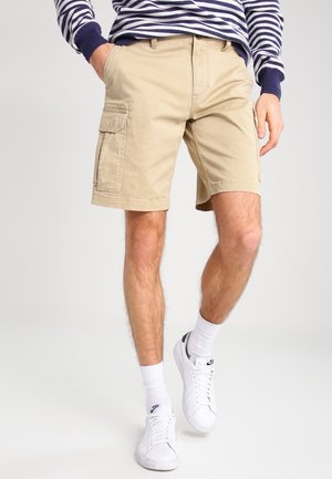Homme portant un short cargo beige, des baskets blanches Nike, des chaussettes blanches montantes et un t-shirt à manches longues rayé bleu marine et blanc, les mains dans les poches.