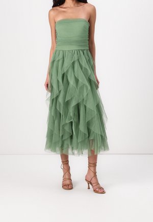 Schulterfreies, gestuftes Midikleid aus weichem grünem Tüll; mit Lagen von Volants, figurbetontem Oberteil und fließendem Rock. Kombiniert mit beigen Riemchenabsätzen.