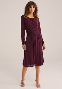 Rochie burgundy cu mâneci lungi, talie strânsă și o strălucire subtilă, căzând sub genunchi. Sandale argintii completează look-ul.