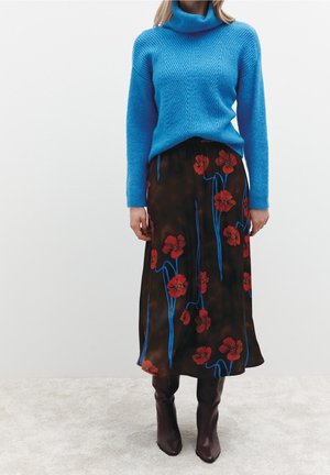 Nainen pukeutunut siniseen pooloneuleeseen, mustaan polvipituiseen hameeseen, jossa on punainen kukkakuosi, sekä tummanruskeisiin polven yli ulottuviin saappaisiin valkoista taustaa vasten.