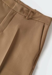 Pantalons beiges ajustés avec passants de ceinture, plis avant et poches latérales, disposés à plat sur une surface blanche.