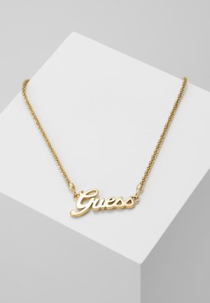Collier chaîne doré avec un pendentif formant le mot « Guess » en écriture cursive, présenté sur une surface géométrique blanche.