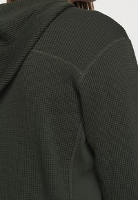 Sudadera con textura en verde oliva y patrón de tejido de gofre, con capucha y costuras visibles a lo largo de las uniones.