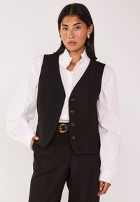 Zwarte wollen vest met een getailleerde pasvorm, voorzien van een V-hals, vier knopen en zijzakken, gecombineerd met een witte ruches-blouse eronder.