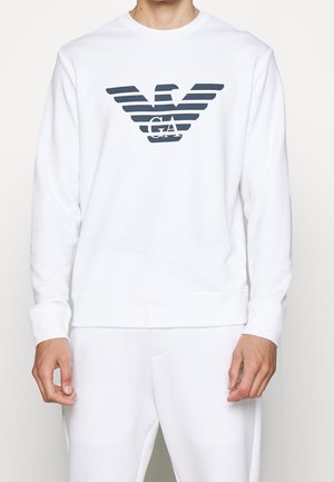 Henkilö pukeutuneena valkoiseen pitkähihaiseen collegepaitaan, jossa suuri tumma Armani-logo rinnan keskellä.