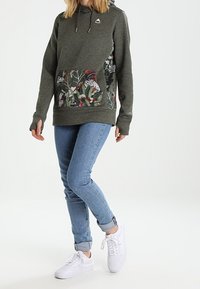 Sudadera de color verde oscuro con un bolsillo frontal estampado, combinada con jeans de color azul claro y zapatillas blancas. Presenta una capucha con cordón y un acento de logo.