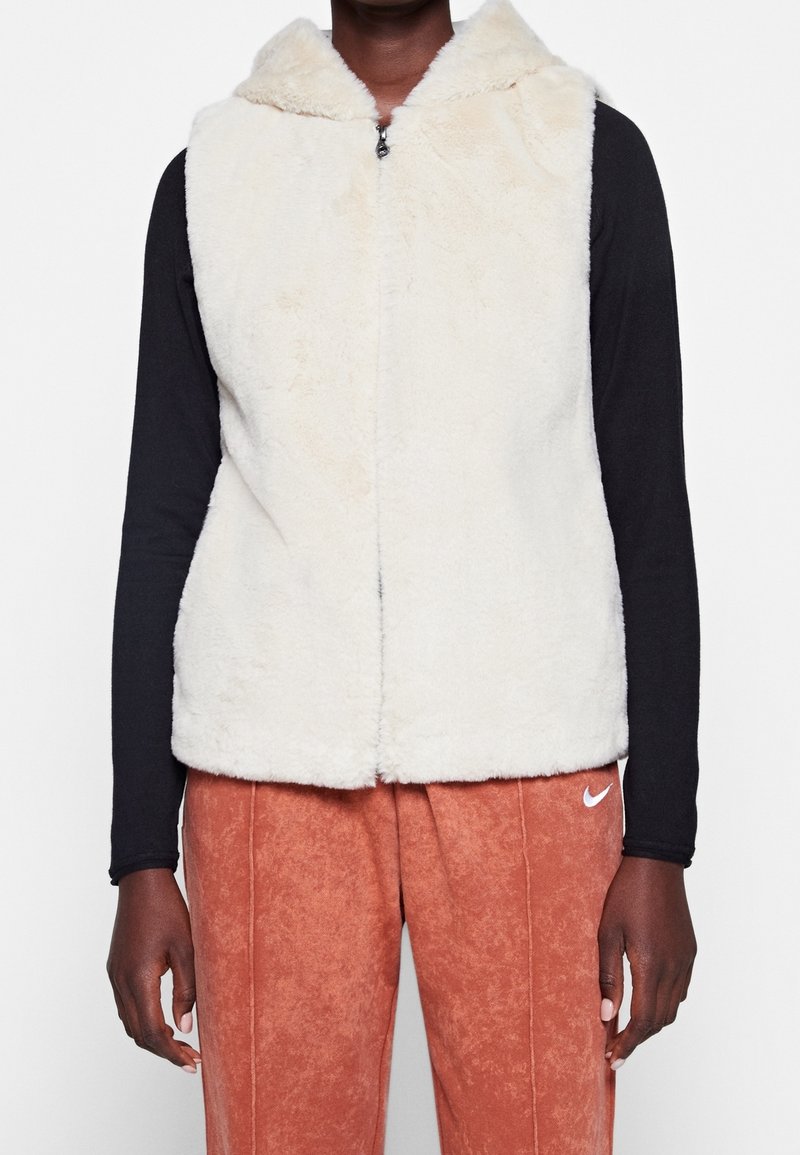 Faux fur krämfärgad väst med huva, dragkedjestängning och utan ärmar, i kombination med texturerade orange byxor. Har en mjuk, fluffig textur.