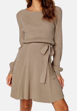 Een vrouw in een beige geribbelde gebreide jurk met lange mouwen en een strikceintuur, staand tegen een effen witte achtergrond.