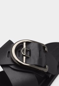 Κοντινή λήψη λουριού μαύρου δερμάτινου σανδαλιού με μεγάλο ασημένιο μεταλλικό αγκράφα και πολλές οπές ρύθμισης.