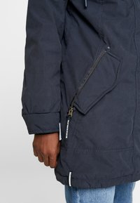 Manche de veste bleu foncé et poche latérale avec fermeture éclair reposant à côté de la main d'une personne portant un jean bleu clair.
