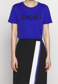 Niebieska koszulka z królewskim odcieniem z czarnym logo "DKNY", zestawiona z czarną spódnicą z białym i niebieskim zamkiem po boku.