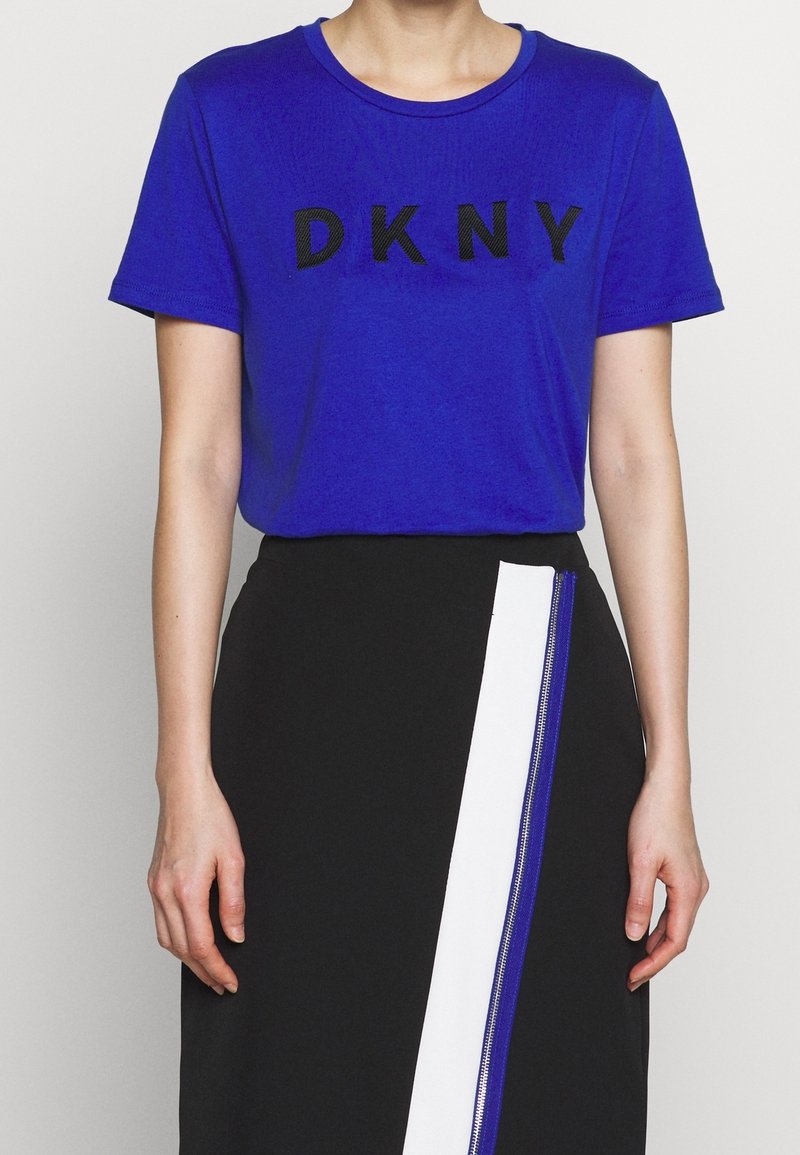 Niebieska koszulka z królewskim odcieniem z czarnym logo "DKNY", zestawiona z czarną spódnicą z białym i niebieskim zamkiem po boku.