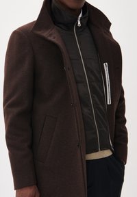 Bruine wollen overjas met een hoge kraag, voorzien van een zwart donsjack eronder, een ritssluiting aan de voorkant en zijzakken.