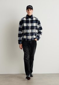Checkered fleece jacket in black, white, and blue, with a zip front and side pockets, paired with dark pants and black shoes.