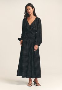 Black long-sleeve dress with a deep V-neck and gathered waist, featuring a white polka dot pattern and flowing skirt.