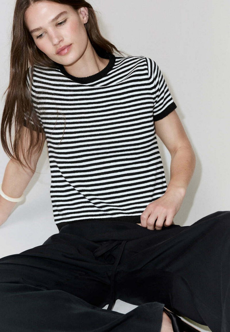 Jeune personne aux longs cheveux bruns portant un haut à manches courtes rayé noir et blanc et un pantalon noir ample, assise devant un fond uni.