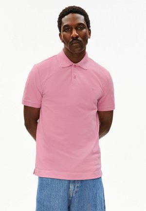 Man met snor, gekleed in een roze poloshirt en blauwe spijkerbroek, staand met handen achter de rug tegen een witte achtergrond.