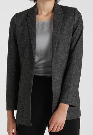Woman wearing dark gray tailored blazer over light gray satin blouse and black pants, standing against plain background.