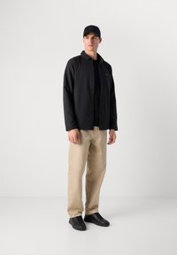 Svart skjortejakke over en svart skjorte, kombinert med beige bukser og svarte sneakers. Jakken har et enkelt design og myk tekstur.