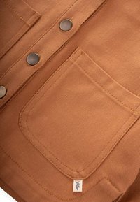 Brun manchester tyg med en ficka, två mörka metallknappar och subtila sömnadsdetaljer. Liten logotags i nedre hörnet.