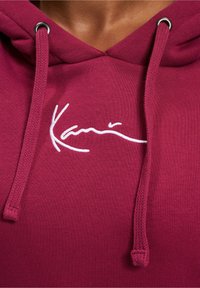 Bordeauxroter Hoodie aus strukturiertem Stoff, mit weißer, kursiver Logostickerei und verstellbarem Kordelzug mit silbernen Ösen.