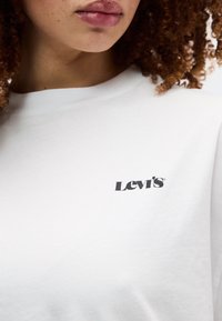 Κοντινό πλάνο ενός ατόμου με σγουρά μαλλιά που φοράει λευκό πουκάμισο Levi's, εμφανίζοντας τα χείλη, το πηγούνι, τον λαιμό και την περιοχή του άνω στήθους.